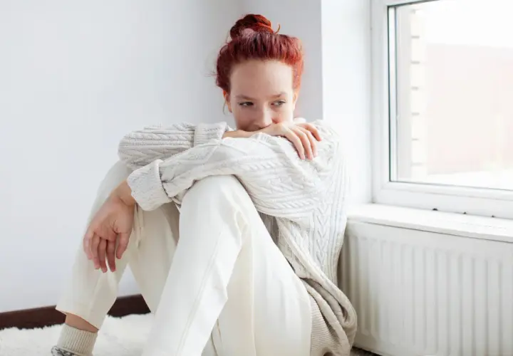 Ragazza con capelli rossi seduta vicino a una finestra, avvolta in un maglione bianco. Potrebbe riflettere su come l'olanzapina a cosa serve per affrontare momenti difficili.
