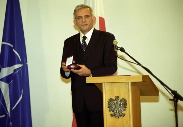 Jerzy Buzek, premier Polski, prezentuje medal. Obok flagi NATO i Polski.