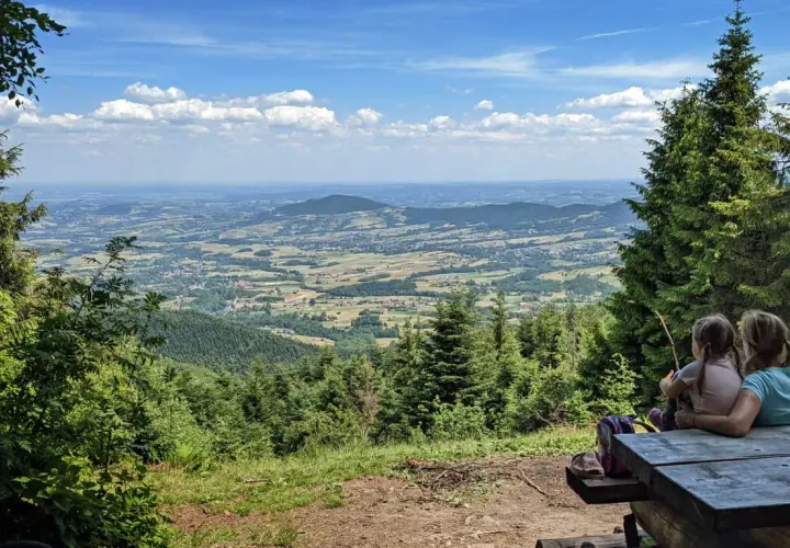 Dwie dziewczynki siedzą przy drewnianym stole, podziwiając rozległą panoramę **Beskidu Makowskiego**. W oddali widać łagodne **szczyty** i malownicze doliny.
