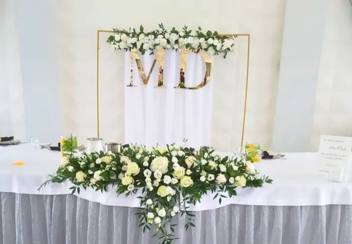 Elegancka dekoracja stołu weselnego z białymi różami i zielenią. Złote litery "MD" na tle białej tkaniny. W tle złota konstrukcja z kwiatami.