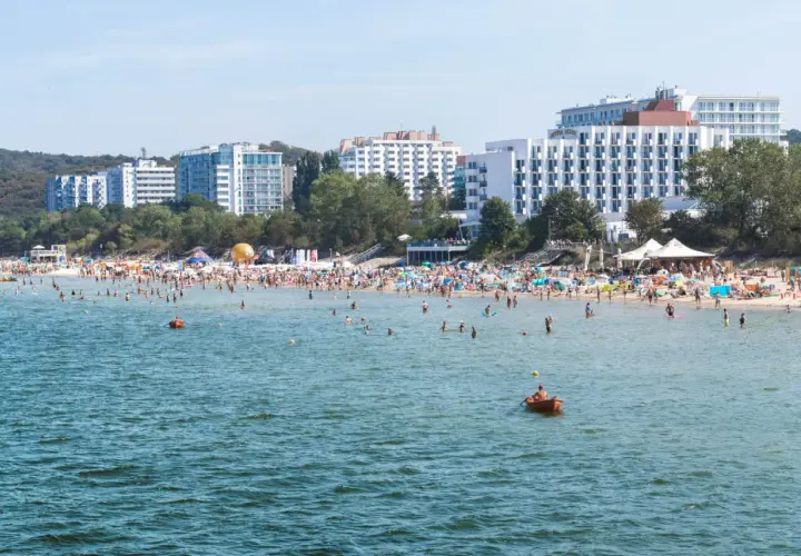 Tłum ludzi na plaży w Międzyzdrojach, kąpielisko pełne plażowiczów i łódek. W tle hotele i drzewa. Międzyzdroje atrakcje w pełnym słońcu.