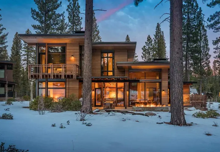 Cozy modern mountain homes with warm interior lights and a roaring fire pit, nestled amongst snow-covered trees.