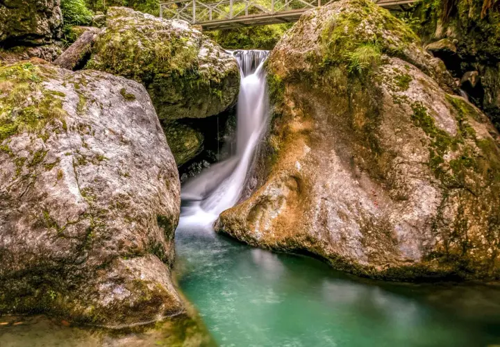 Cascada en las gargantas de Kakueta, con agua turquesa y rocas cubiertas de musgo. Un puente metálico cruza sobre la caída de agua.