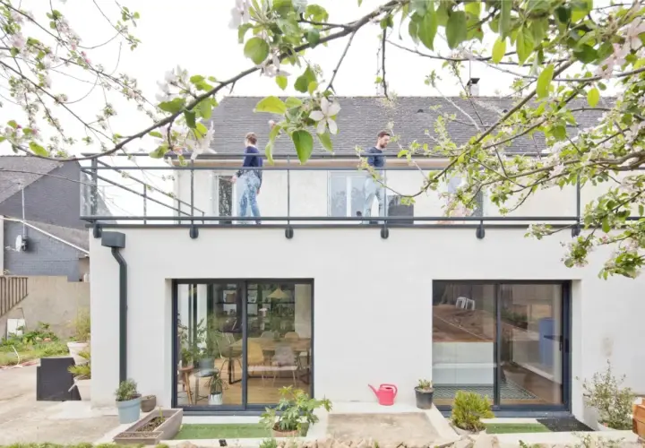 Surélévation maison 50m2 : deux personnes sur un balcon moderne, sous des branches fleuries. Le prix de ce projet est attractif.
