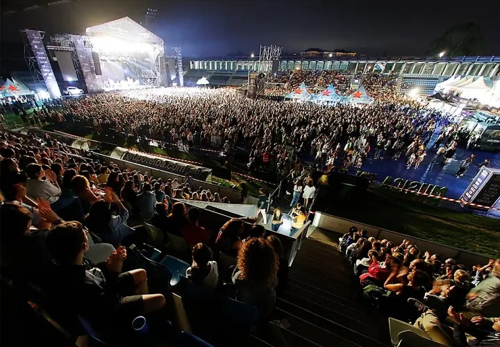 Multitud disfrutando de conciertos Torrelavega hoy. Escenario iluminado, miles de personas vibran con la música en un estadio.