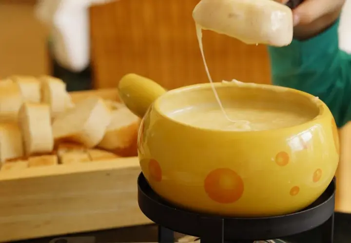 Un pezzo di pane viene intinto in una fonduta di pecorino fumante, servita in una ciotola gialla decorata.