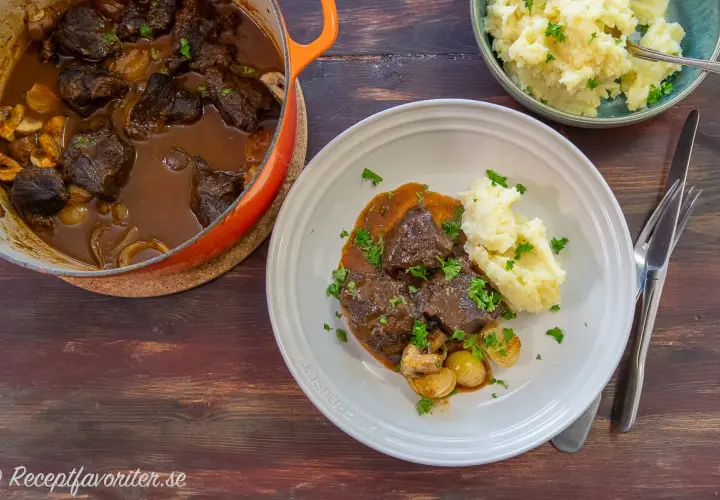 A Dutch oven filled with braised beef and mushrooms, next to a plate of beef with mashed potatoes and a bowl of mashed potatoes.