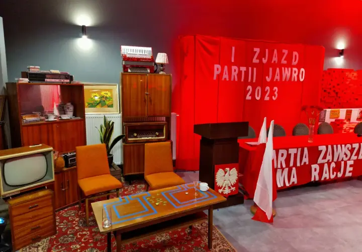 Scena imprezy w stylu PRL: meble, radio, telewizor, akordeon, czerwony transparent z napisem.