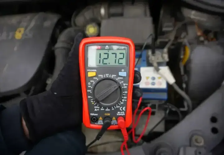 Ręka w czarnej rękawiczce trzyma miernik napięcia 12v, pokazujący 12.72V, przy akumulatorze samochodowym.