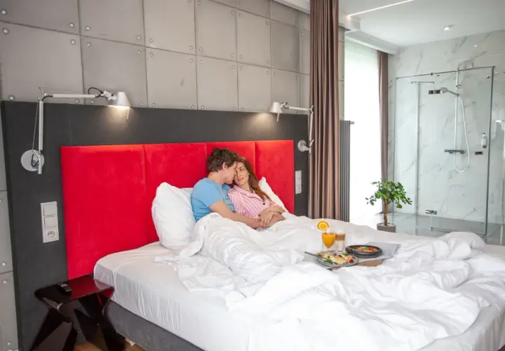 Para w łóżku z tacą śniadaniową, w nowoczesnym pokoju hotelowym z prysznicem.