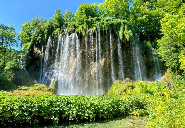 A stunning waterfall cascades down a rocky cliff, surrounded by lush green foliage. Perfect for Croatia hiking trips, this scene captures the natural beauty of Plitvice Lakes National Park.