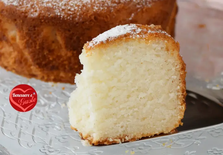 Una fetta di torta senza glutine, soffice e dorata, cosparsa di zucchero a velo, su un piatto trasparente.