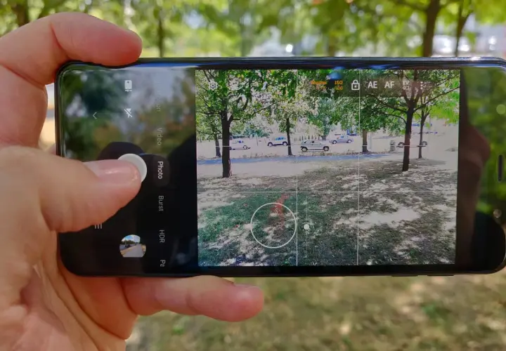 Dłoń trzyma smartfon, na ekranie którego widać interfejs aparatu. Aplikacja aparatu jest gotowa do zrobienia zdjęcia.