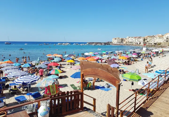 Plage animée à Cefalù en Sicile, avec des parasols colorés, des baigneurs dans la mer bleue et des bâtiments historiques en arrière-plan.