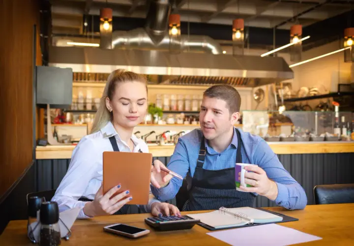Para omawia koszty uzyskania przychodu w restauracji, analizując dane na tablecie i kalkulatorze.