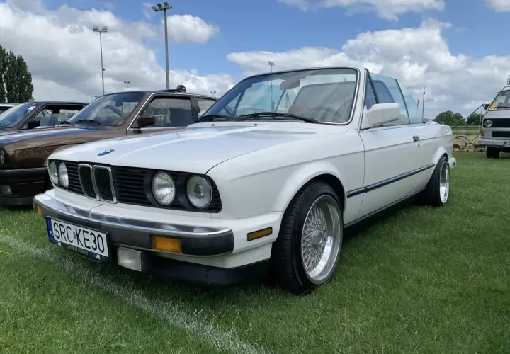 Białe BMW 325i kabriolet z chromowanymi felgami stoi na trawie pod błękitnym niebem z chmurami.
