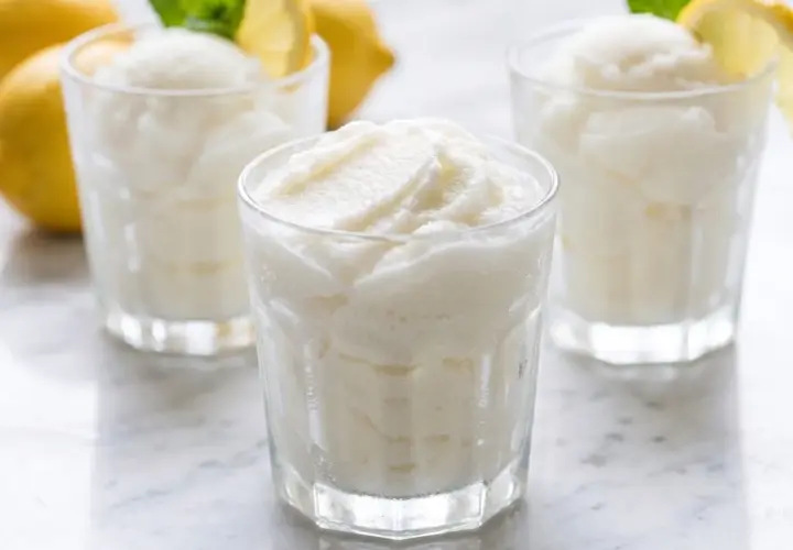Tres vasos de sorbete de limón refrescante, con limones amarillos y hojas de menta en el fondo.