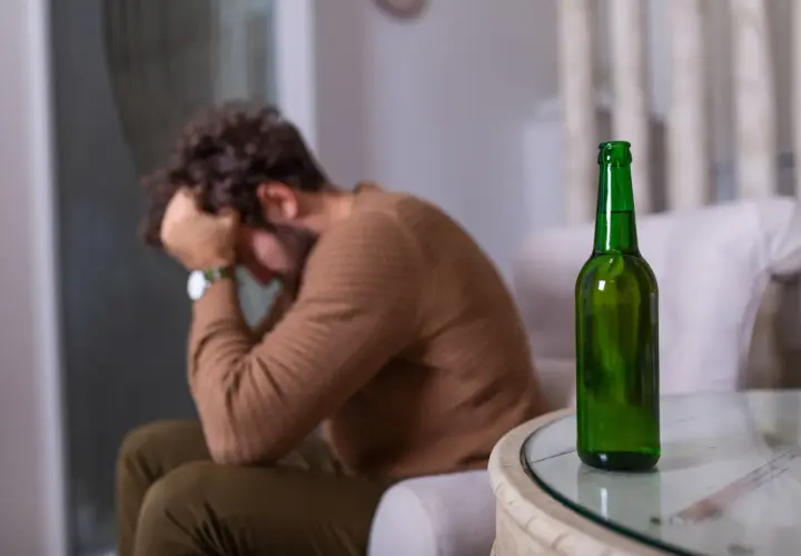 Mężczyzna z głową w dłoniach siedzi obok pustej butelki piwa. Może to symbolizować uczulenie na alkohol lub jego nadużywanie.