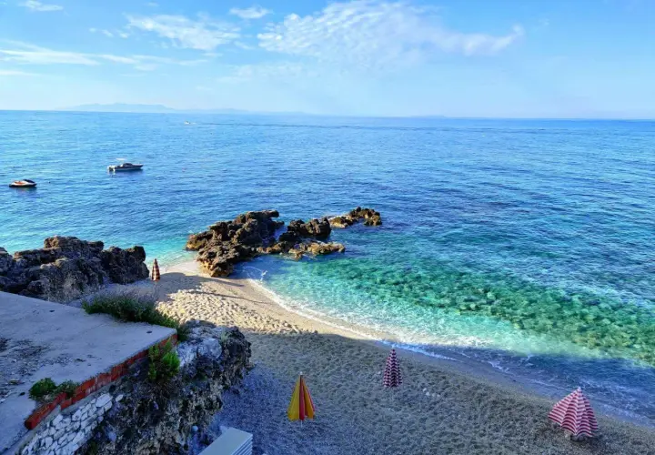 Albania, czerwiec: słoneczna pogoda, turkusowe morze, kamienista plaża z parasolami i łódkami na horyzoncie.