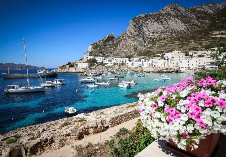 Vue d'un port pittoresque en Italie, avec des bateaux dans des eaux turquoise et des maisons blanches accrochées à flanc de colline. Un bouquet de fleurs roses et blanches encadre la scène, évoquant la beauté de Marsala, Italie.