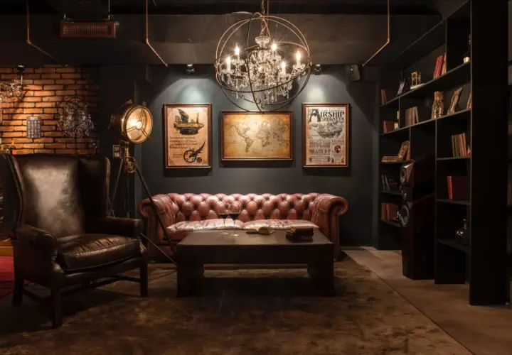 Sofá de cuero, sillón y mapa del mundo en una sala con decoración steampunk. Lámpara de pie industrial y estanterías llenas de libros.