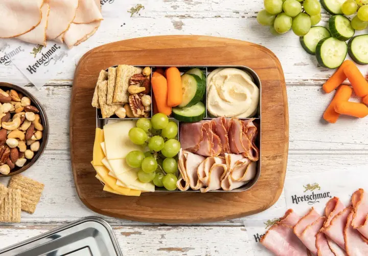 A bento box with cheese, grapes, crackers, nuts, hummus, carrots, and cucumber. Perfect for when are bento boxes eaten cold.