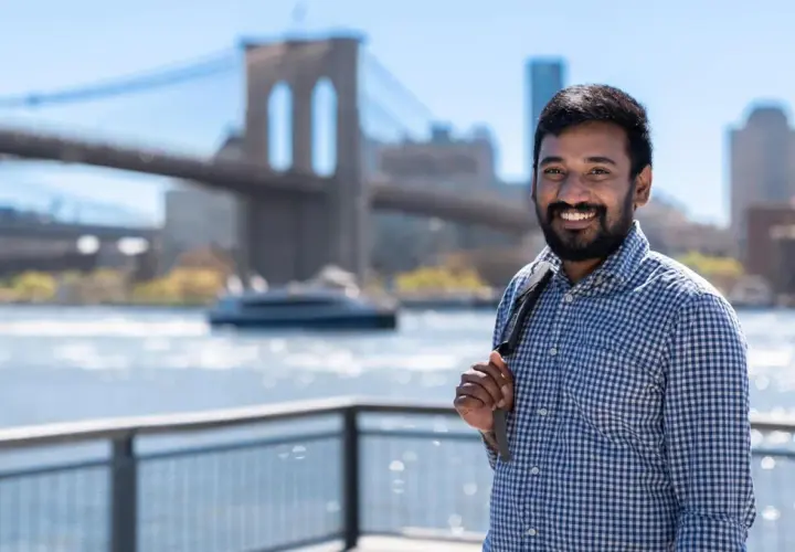 A smiling man with a beard, wearing a blue checkered shirt, stands with the Brooklyn Bridge in the background. He's a student in the Pace University MPA program.