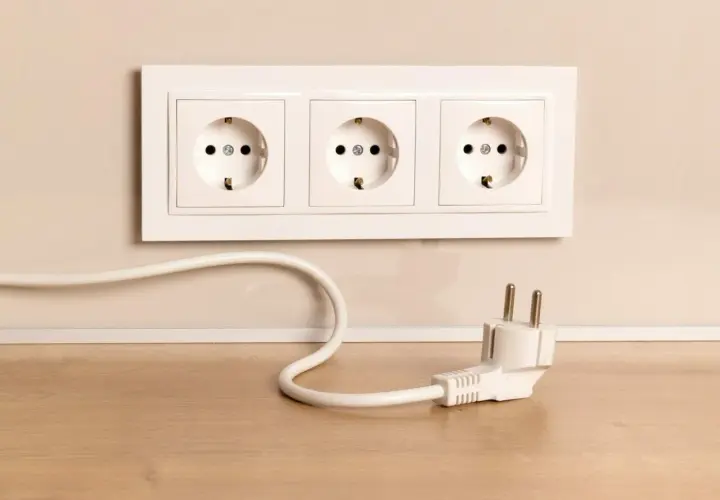 Biały kabel z wtyczką leży na drewnianym blacie, obok gniazdek elektrycznych w ścianie.