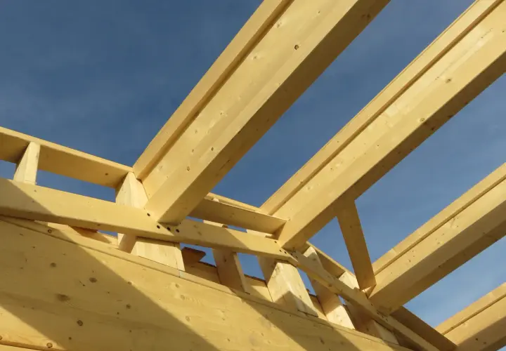 Structure en bois d'une charpente fermette sous un ciel bleu. Les poutres de bois clair forment un motif géométrique complexe.