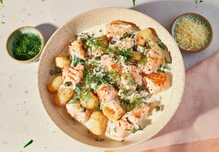 Pyszne gnocchi z łososiem w kremowym sosie, udekorowane koperkiem i kaparami. Obok miseczka z tartym parmezanem.