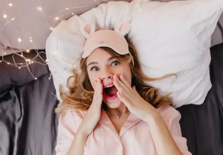 En tjej med sovmask och rosa pyjamas ligger i sängen, redo för att sova med sminket kvar. Hon ser överraskad ut.