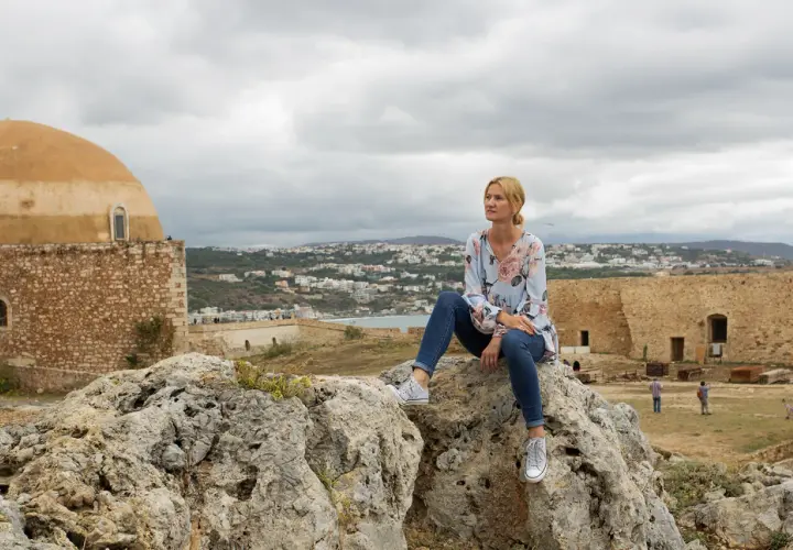 Kobieta siedzi na skale, podziwiając widok miasta. Kreta pogoda październik, chmury na niebie.