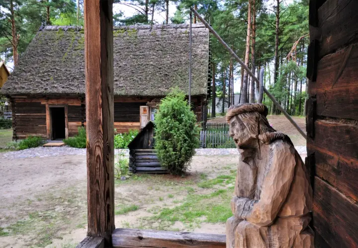 Drewniana rzeźba postaci, tradycyjna chata z mchem na dachu i zielone drzewa tworzą klimatyczne **kadzidło atrakcje** w skansenie.