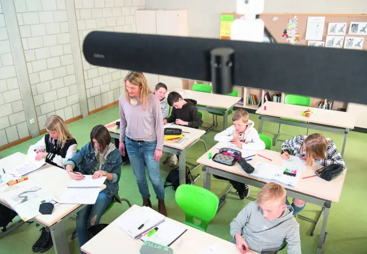 Lehrerin betreut Schüler im Klassenzimmer, die an ihren Tischen arbeiten. Ein modernes Setup für hybriden Unterricht.