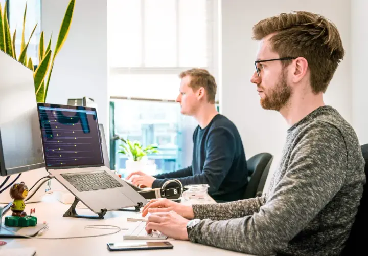 Dwóch programistów pracuje przy komputerach. Jeden z nich ma okulary, co może sugerować refundację okularów przez pracodawcę.