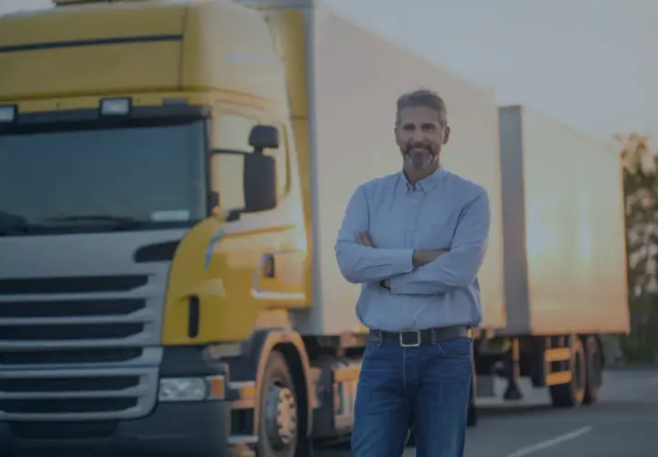 Uśmiechnięty kierowca ciężarówki złożył ręce na piersi. Obok stoi żółta ciężarówka. OCP co to? To jego praca.