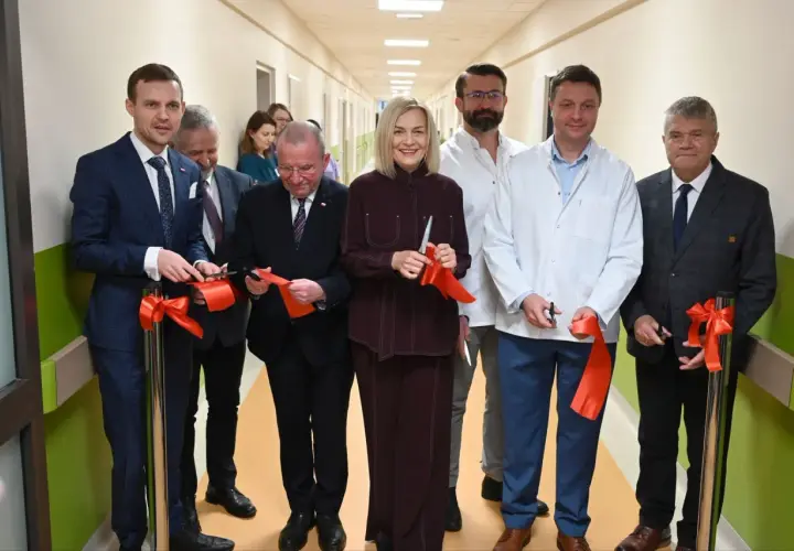 Grupa osób w garniturach i kitlach przecina czerwone wstęgi w szpitalnym korytarzu.