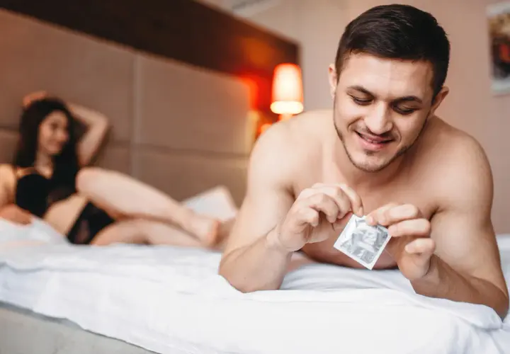 Mężczyzna w łóżku przygotowuje się do intymnego spotkania, trzymając w dłoni prezerwatywę. Kobieta leży w tle. Rozmiary prezerwatyw są kluczowe dla komfortu.