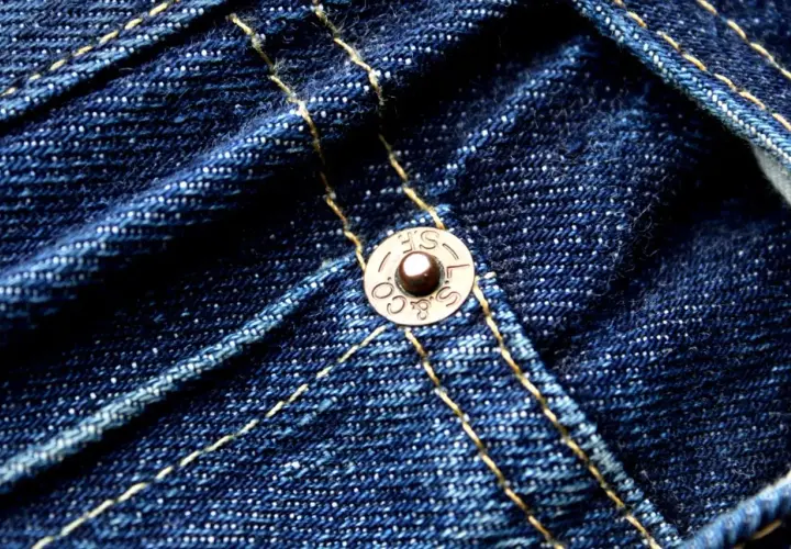 Close-up of dark blue denim with yellow stitching. A copper rivet, marked "S.F. & CO. L.S.", is visible, a testament to the durability of these jeans.