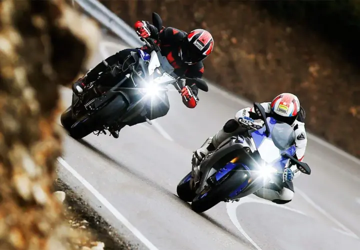 Dos pilotos compiten en una carretera sinuosa, uno en una Yamaha mas rápida, inclinándose en una curva cerrada.