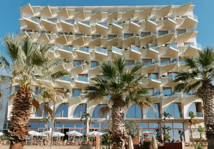 Nowoczesny hotel z białymi, geometrycznymi balkonami i palmami przed fasadą. Epidamn white sensation.