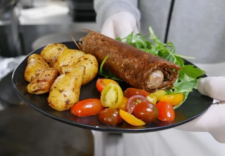 Une galette saucisse roulée, servie avec des pommes de terre rôties et des tomates cerises colorées, sur une assiette noire.