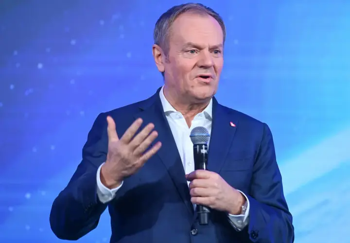 Donald Tusk w garniturze, przemawiający do mikrofonu na tle niebieskiego ekranu.