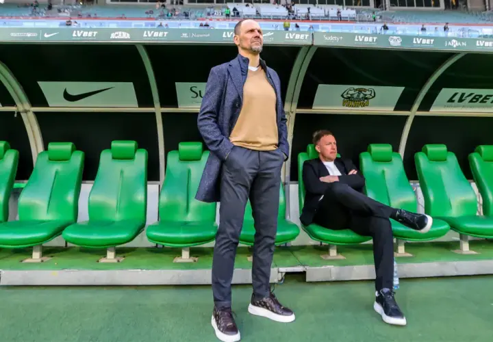 Trener Śląska Wrocław stoi na tle zielonych siedzeń. Obok siedzi inny mężczyzna.