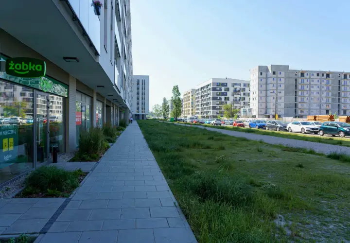 Nowoczesne osiedle w Ursusie Centralnym. Chodnik prowadzi do sklepu Żabka, a dalej widać parking z samochodami i budynki mieszkalne.
