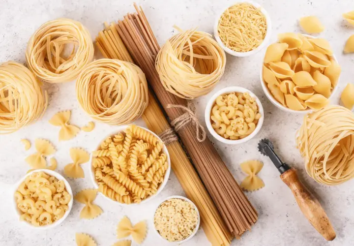 Różnorodne rodzaje makaronu: tagliatelle, fusilli, muszelki, gwiazdki, farfalle i inne, ułożone na jasnym tle.