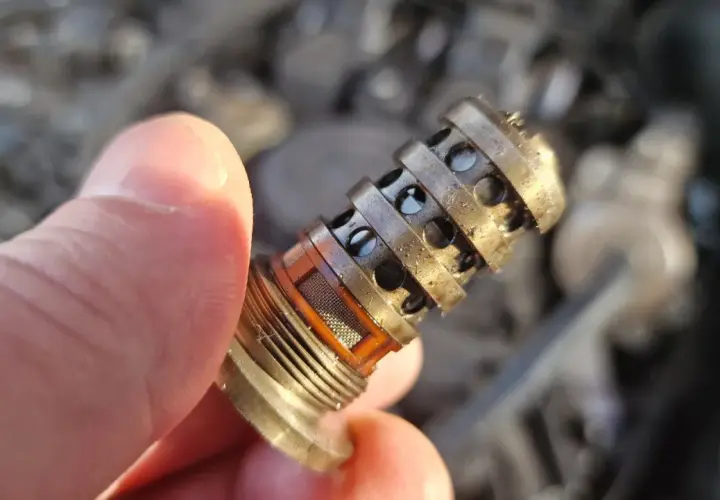Uszkodzony zawór zmiennych faz rozrządu objawy: brudny, metalowy element z otworami, trzymany w palcach, z widocznym filtrem.