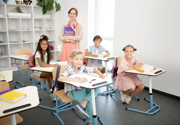 Uśmiechnięta nauczycielka z książką "English grammar" i dzieci przy ławkach. Widać, że to świetny moment, kiedy najlepiej się uczyć.