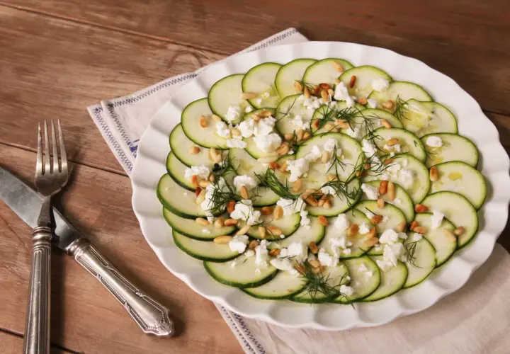 Delicioso carpaccio de calabacín con queso feta desmenuzado, piñones tostados y eneldo fresco, servido en un plato blanco sobre una mesa de madera.