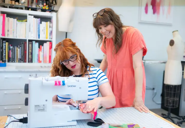 Two women are focused on sewing seams on a white Brother sewing machine in a bright studio.